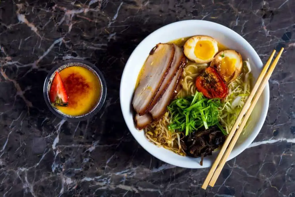 A bowl of ramen on a marble table next to a dessert