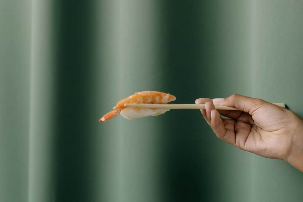 A person off-screen holding a piece of shrimp nigiri with chopsticks