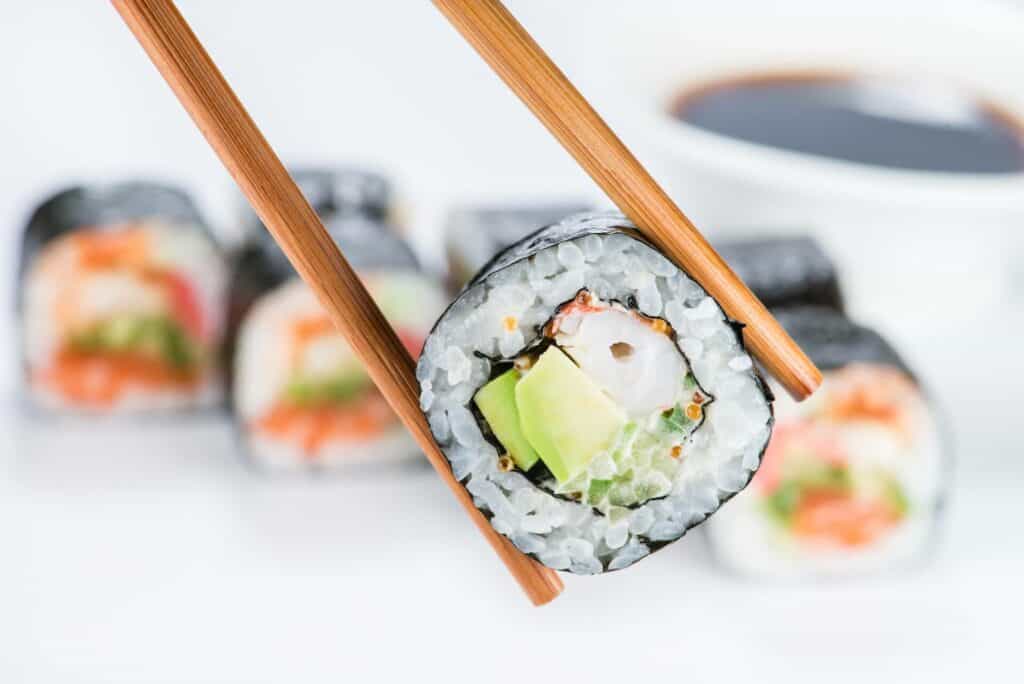 Piece of sushi held by chopsticks in front of other pieces on a plate  